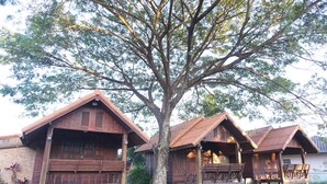 A row of wooden cabins with sloped roofs, surrounded by a large tree and a brick wall. - Chanprasert House (Udon Thani)