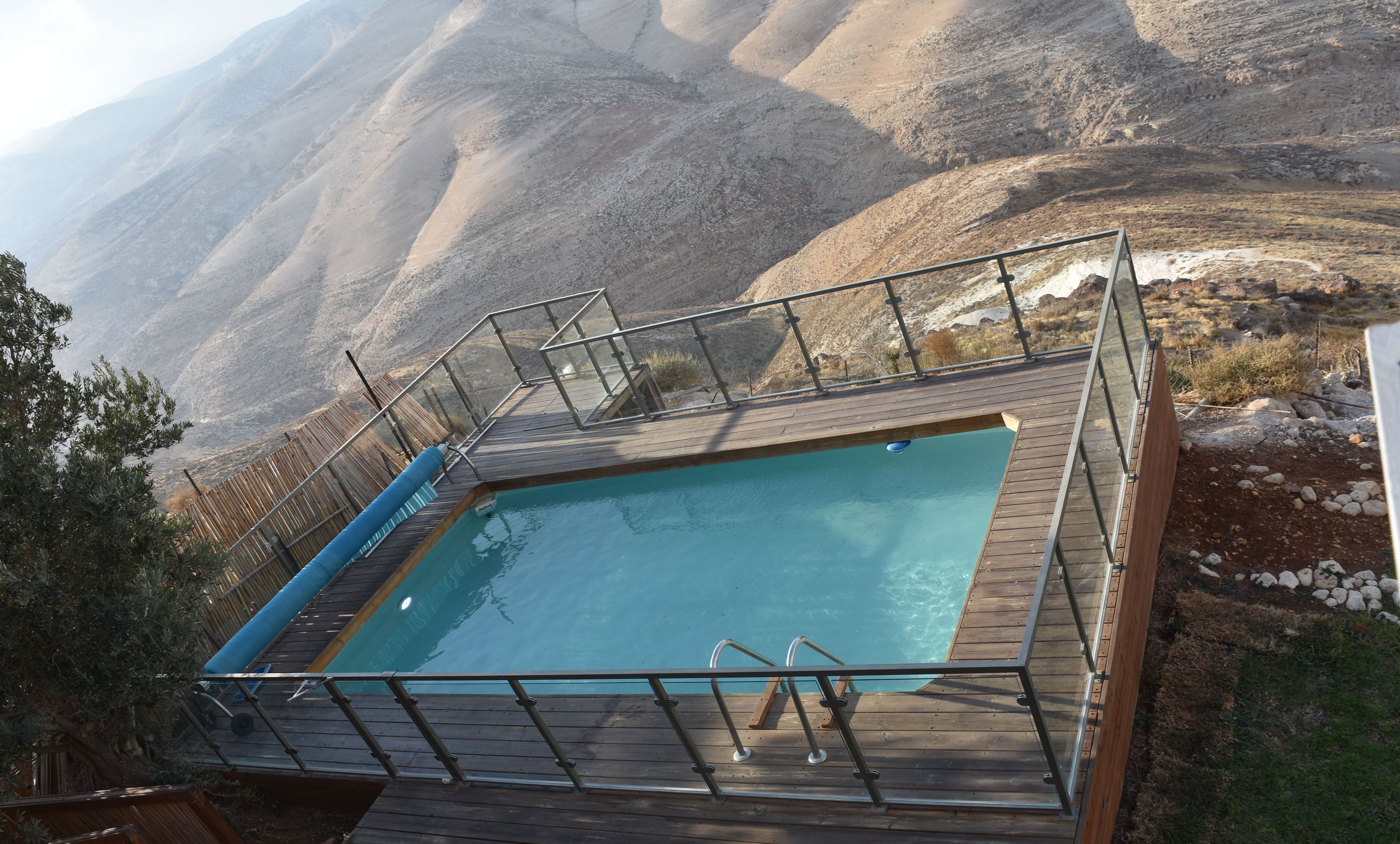 A rectangular pool with a wooden deck and metal railings, surrounded by rocky terrain and sparse vegetation.