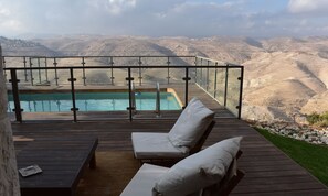 Terraza de madera con barandilla de vidrio que da a una piscina y un paisaje montañoso.