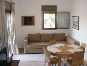 A living room with a beige sofa, a wooden dining table with four chairs, and two windows with curtains.