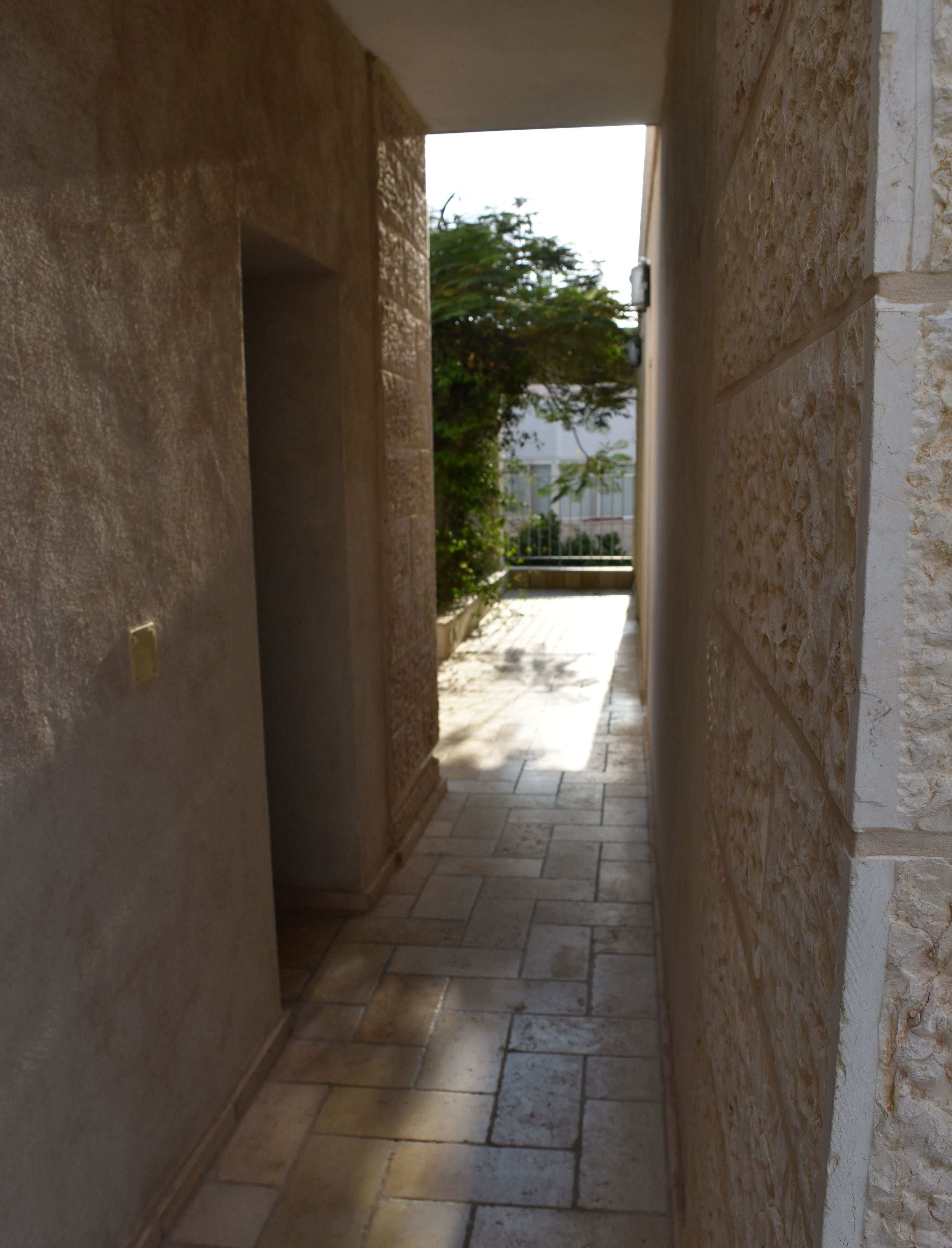 A narrow corridor with stone walls and a tiled floor, leading to an outdoor area with a tree and a fence.