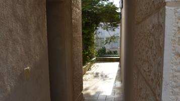 A narrow corridor with stone walls and a tiled floor, leading to an outdoor area with a tree and a fence.