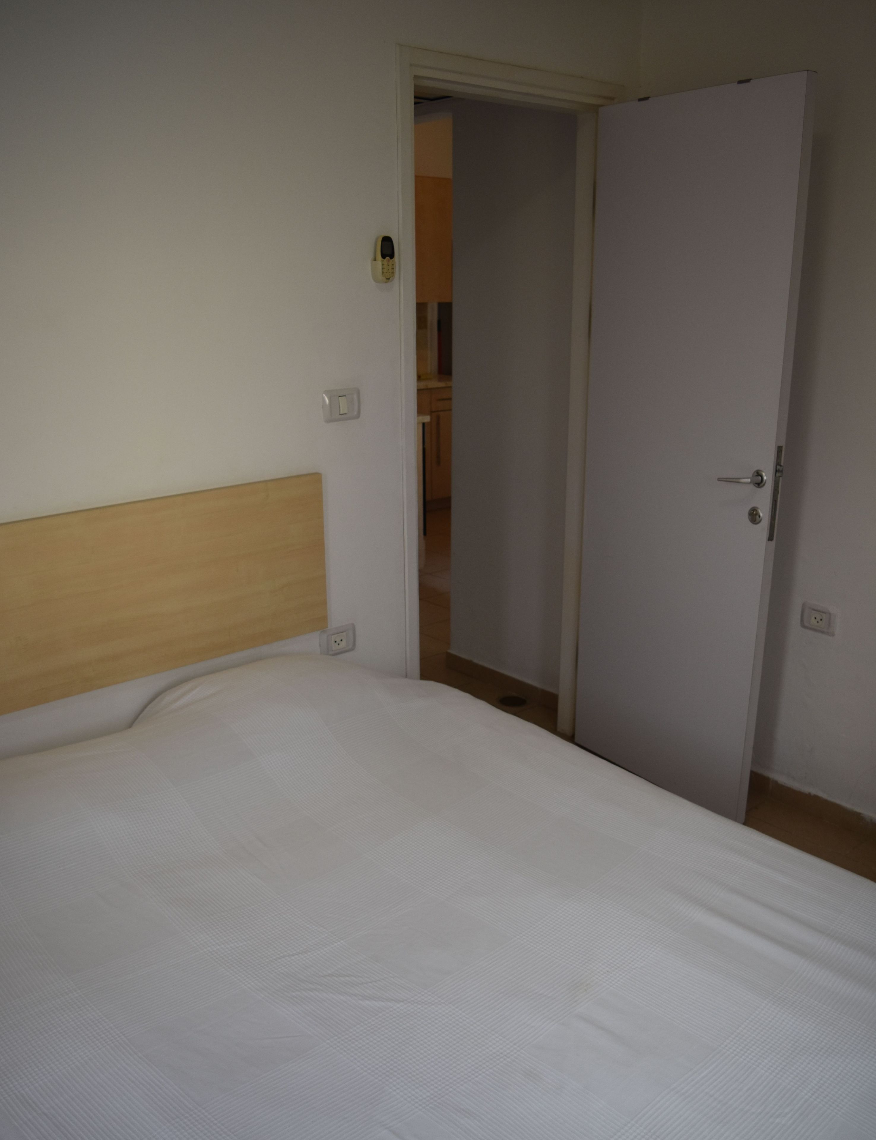A bedroom with a bed, a wooden headboard, a white door, and a visible bathroom in the background.