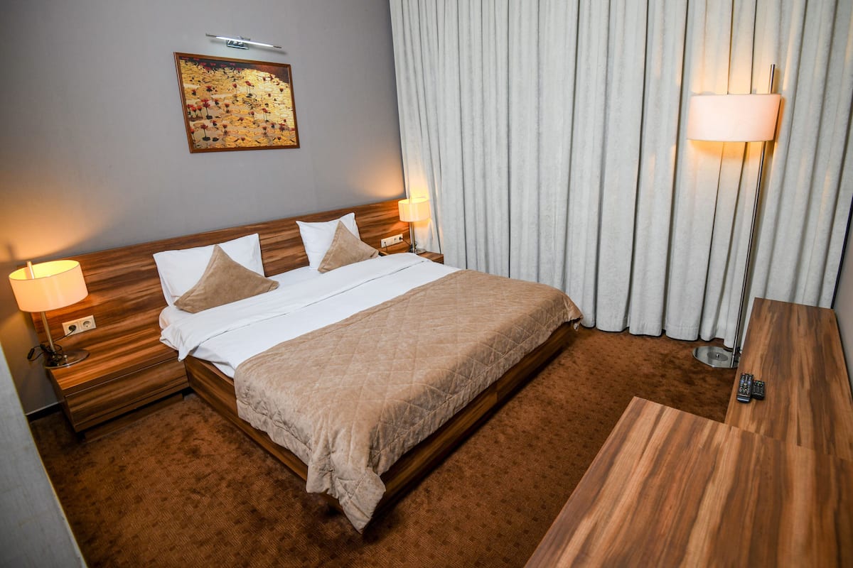 A hotel room with a large bed, two bedside lamps, a wooden headboard, a painting on the wall, and a curtain.