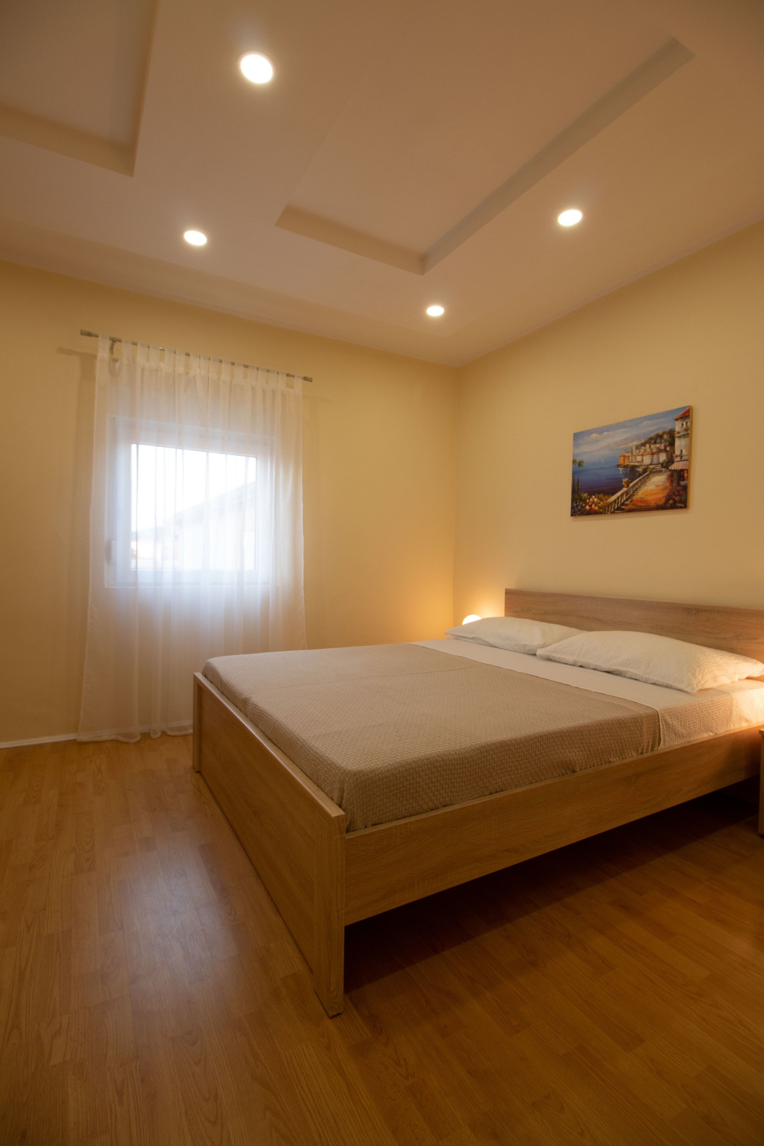 A bedroom with a wooden bed, a painting on the wall, and a window with sheer curtains.