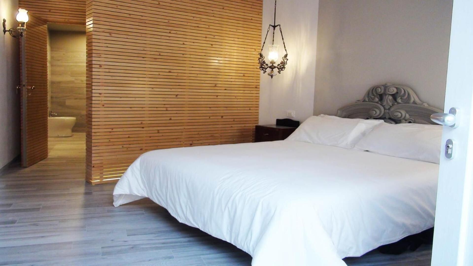 A modern bedroom with a wooden slatted wall, a large bed with white linens, a decorative headboard, and a hanging light fixture.