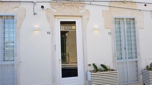 Ein Hotel Eingang mit einem Schild, auf dem 'Room 55' steht und der Zahl '53' an der Wand. Zwei Kübelpflanzen stehen auf dem Boden.