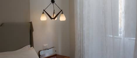 Una habitación con cama, una mesita de noche con espejo, una lámpara colgante y una ventana con cortinas translúcidas.