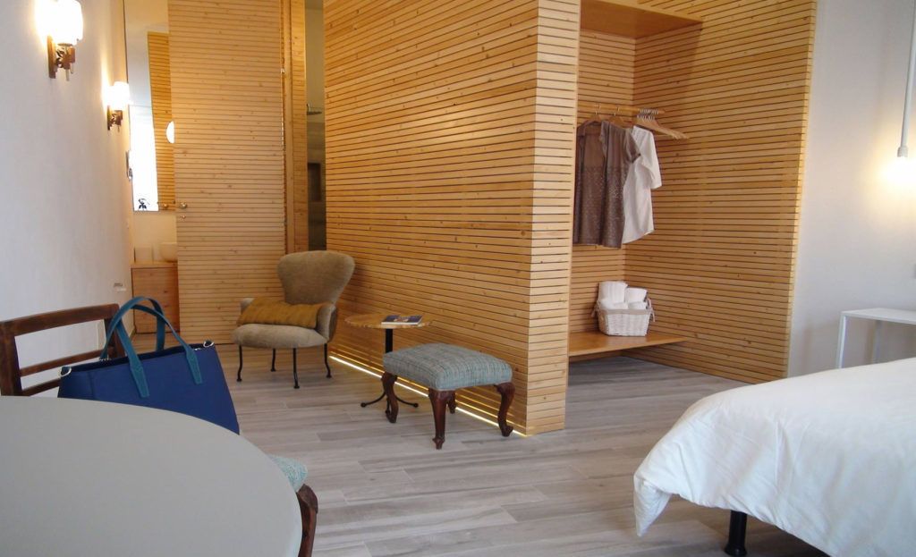 A hotel room with a wooden partition, a chair, a small table, a bench, a bed, and a wardrobe with hanging clothes.
