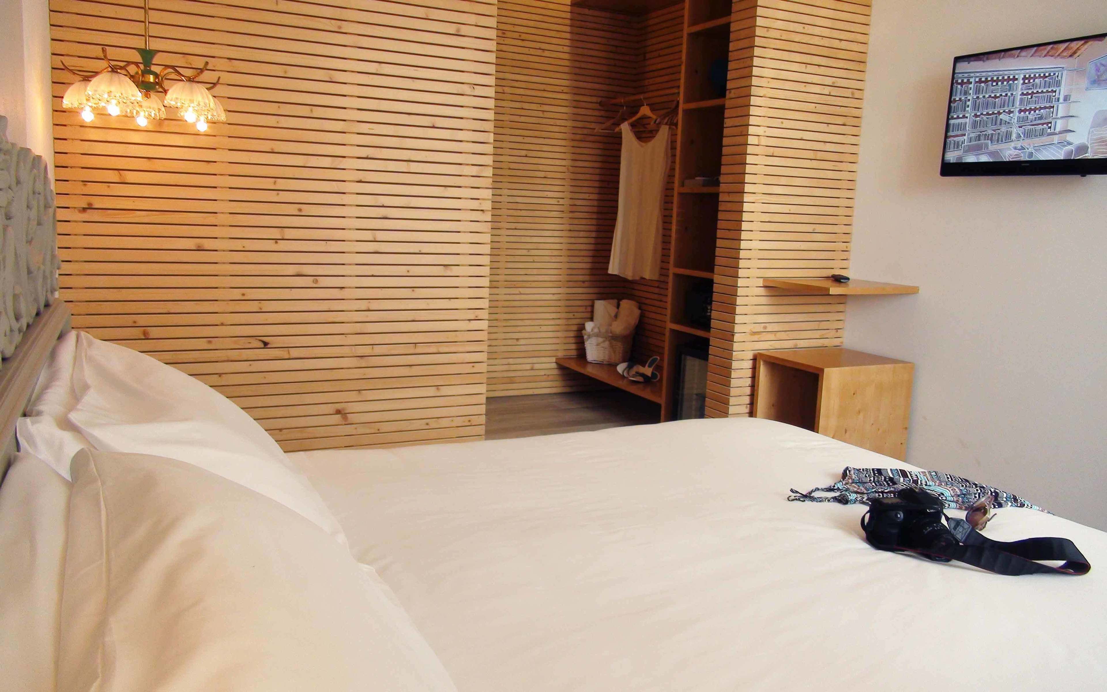 Une chambre avec un mur recouvert de lambris en bois, un lit avec du linge de lit blanc, un luminaire suspendu, une armoire en bois et une petite table en bois.