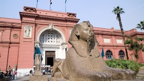 A large stone sphinx statue in front of a pink building with arched windows and flags. - Nile plaza hostel (Cairo)