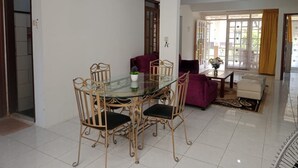 A dining area with a glass table and metal chairs, a purple sofa, and a white armchair. - Villa Kota Bunga Anggrek (Cipanas)