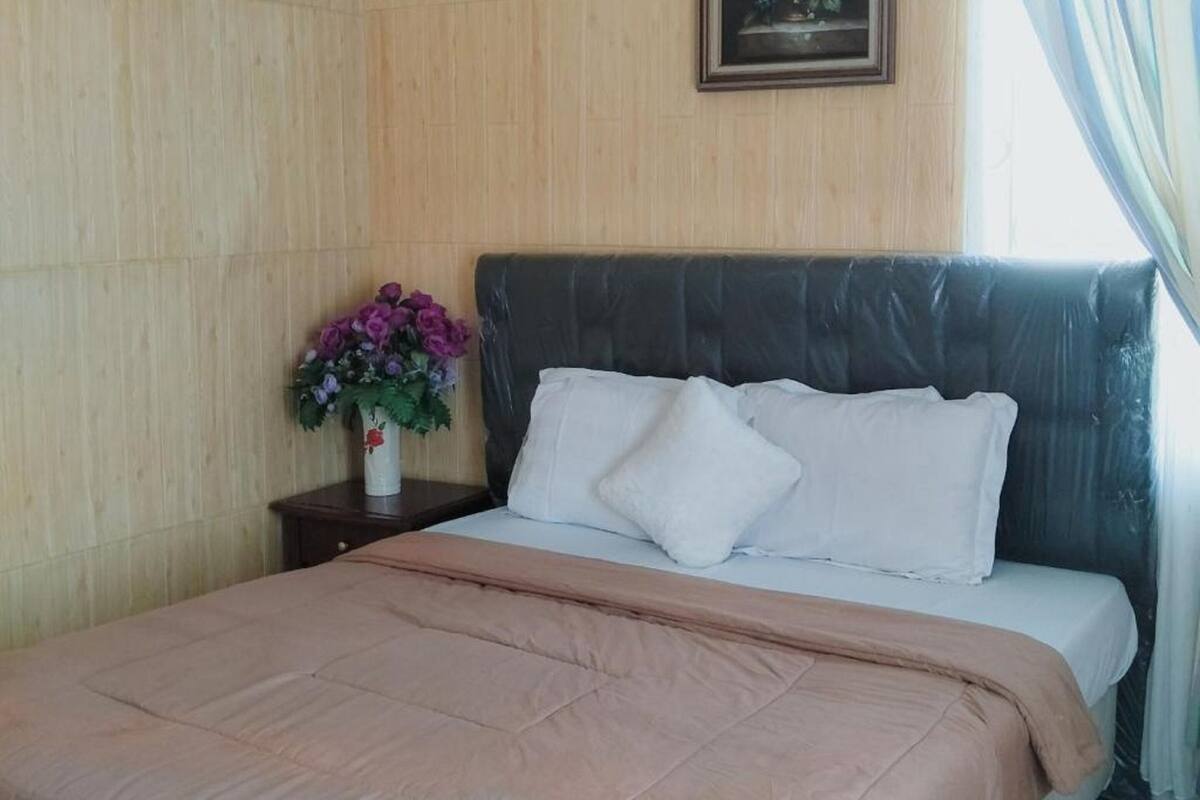 Ein ordentlich bezogenes Bett mit einer rosafarbenen Decke, einem weißen Kopfkissen und einem Blumenstrauß aus violetten Blüten auf einem Nachttisch aus Holz.