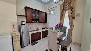 A modern kitchen with dark wooden cabinets, a stainless steel refrigerator, and a dining area with a glass table and two chairs. - Villa Kota Bunga Anggrek (Cipanas)