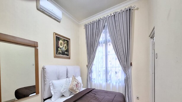 A bedroom with a bed, a mirror, a painting, curtains, and a radiator. - Villa Kota Bunga Anggrek (Cipanas)