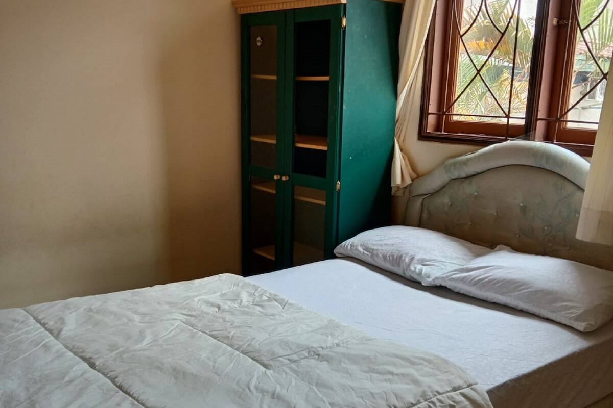 A single-bed room with a green wardrobe, a window with curtains, and a bed with white bedding.