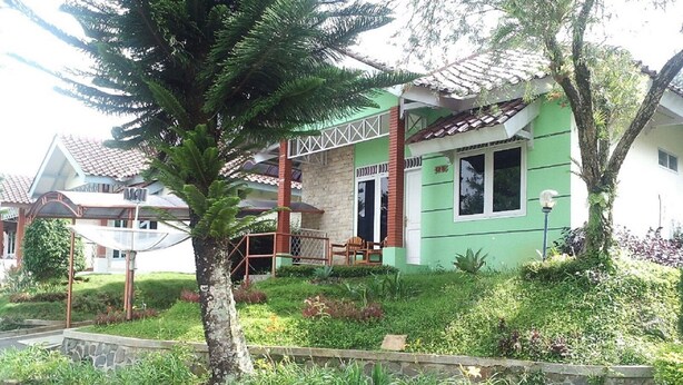 A green house with a tiled roof, a porch, and a garden with various plants. - Villa Kota Bunga Cosmos (Cipanas)