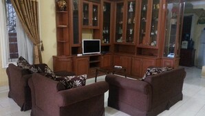 A living room with a sectional sofa, a TV on a stand, and a wooden cabinet with glass doors. - Villa Kota Bunga Cosmos (Cipanas)