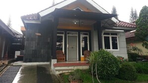 A two-story house with a front porch, a small garden, and a visible garage door. - Villa Kota Bunga Cosmos (Cipanas)