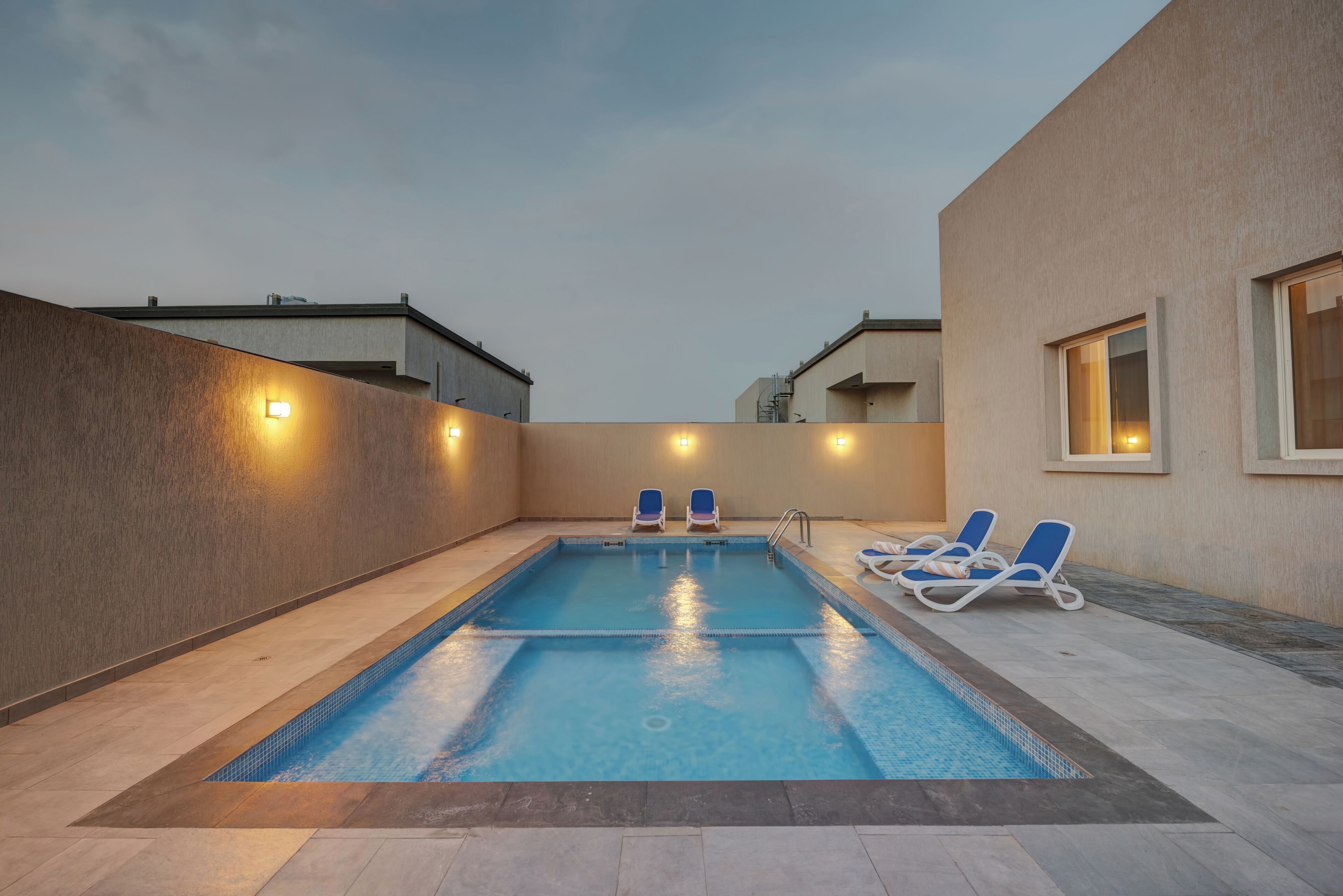 A rectangular swimming pool with two blue lounge chairs on one side and a wall with two windows on the other.