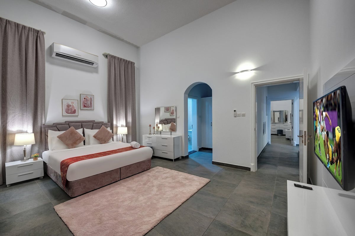 Una habitación de hotel moderna con una cama grande, una mesita de noche con lámpara, aire acondicionado, televisión y vistas al pasillo.