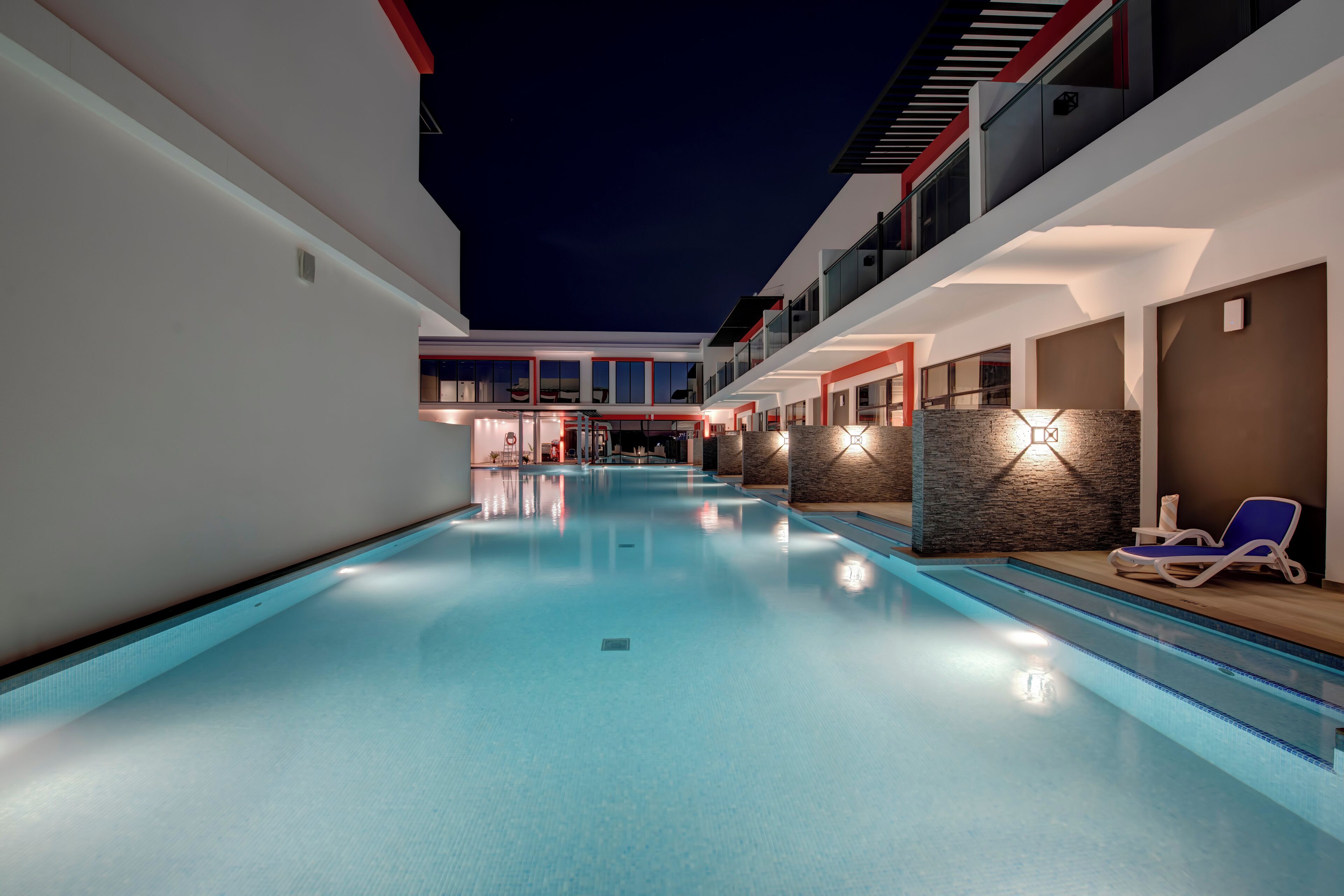 A modern hotel pool area with a clear pool, lounge chairs, and a staircase leading to the upper floors.