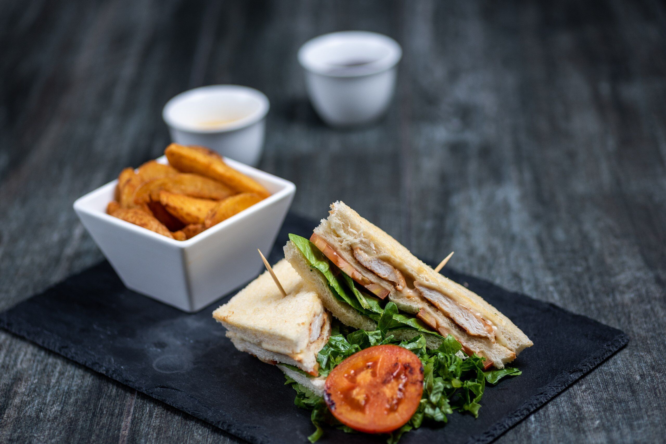 A sandwich with lettuce and tomato on a slate plate, accompanied by a bowl of fries and two small containers of sauce.