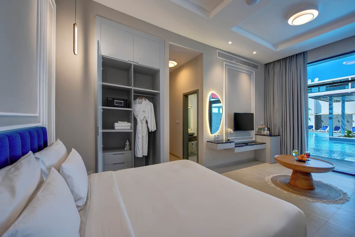 Habitación de hotel moderna con una cama grande, vestidor, televisión, mesa de comedor con fruta y vistas a una piscina y balcón.