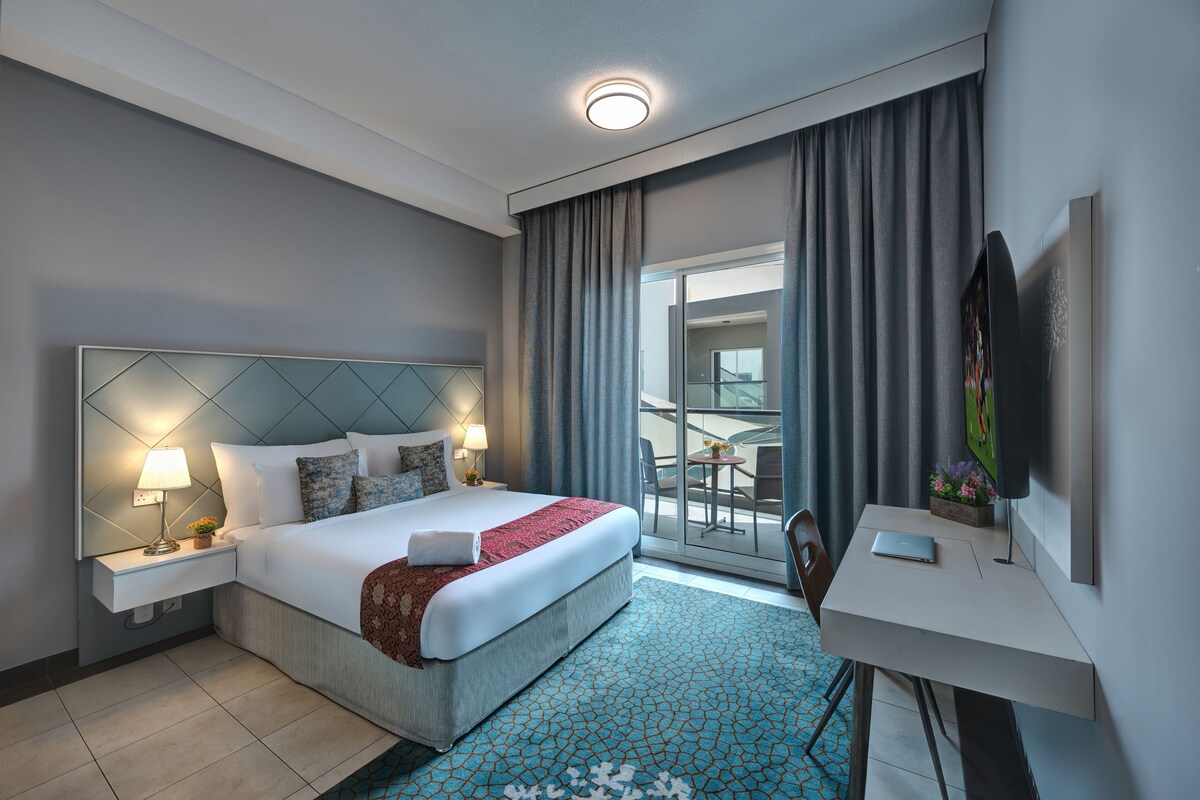 A modern hotel room with a bed, bedside table, lamp, and a desk with a laptop.