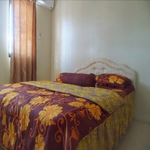 Una cama con una colcha estampada con motivos florales y dos almohadas, cabecera blanca y una ventana con cortinas.