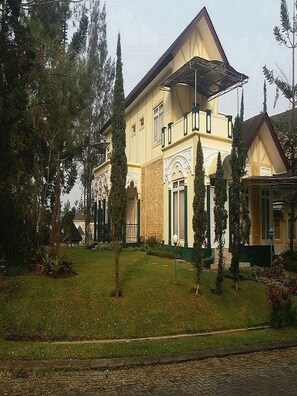 A two-story building with a symmetrical facade, a central entrance with a decorative arch, and a small garden with trimmed grass and a few trees. - Villa Kota Bunga Protea (Cipanas)