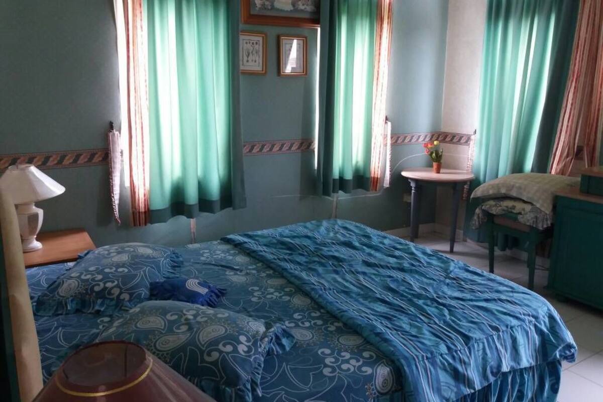 A bedroom with a bed, a bedside table, a chair, a small table with a vase, and curtains.