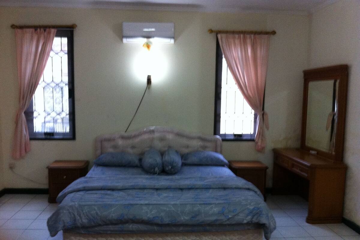 A bedroom with a bed, two nightstands, a dresser, a mirror, and two windows with curtains.