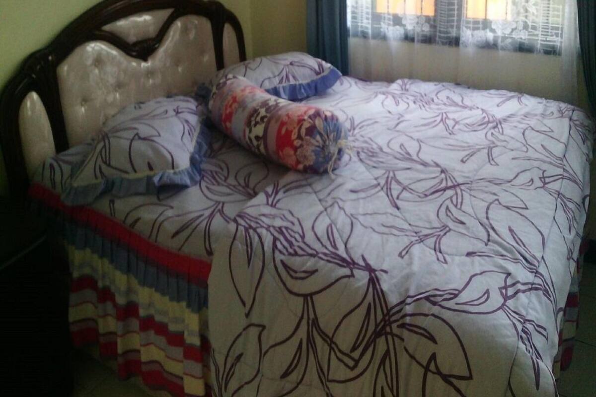 A bedroom with a bed, a wooden headboard, a window with a metal grill, and curtains.