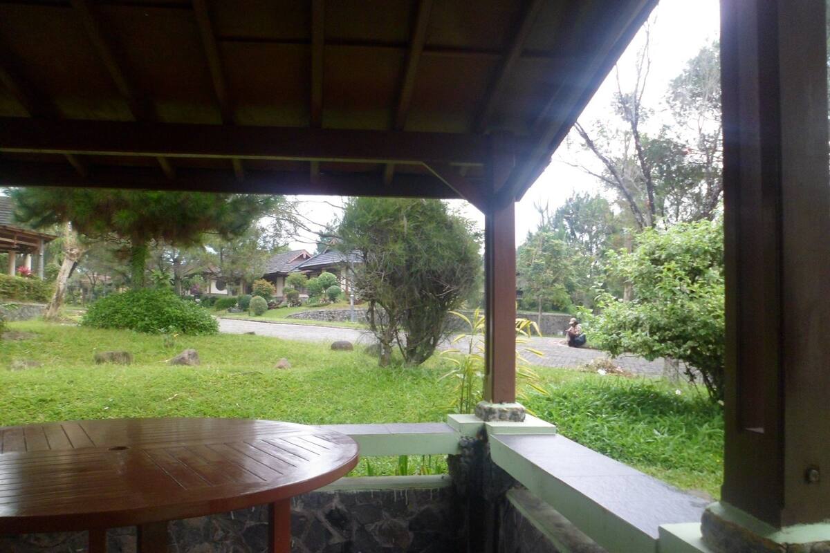 A covered outdoor area with a wooden table, stone wall, and a view of a garden and a house in the background.