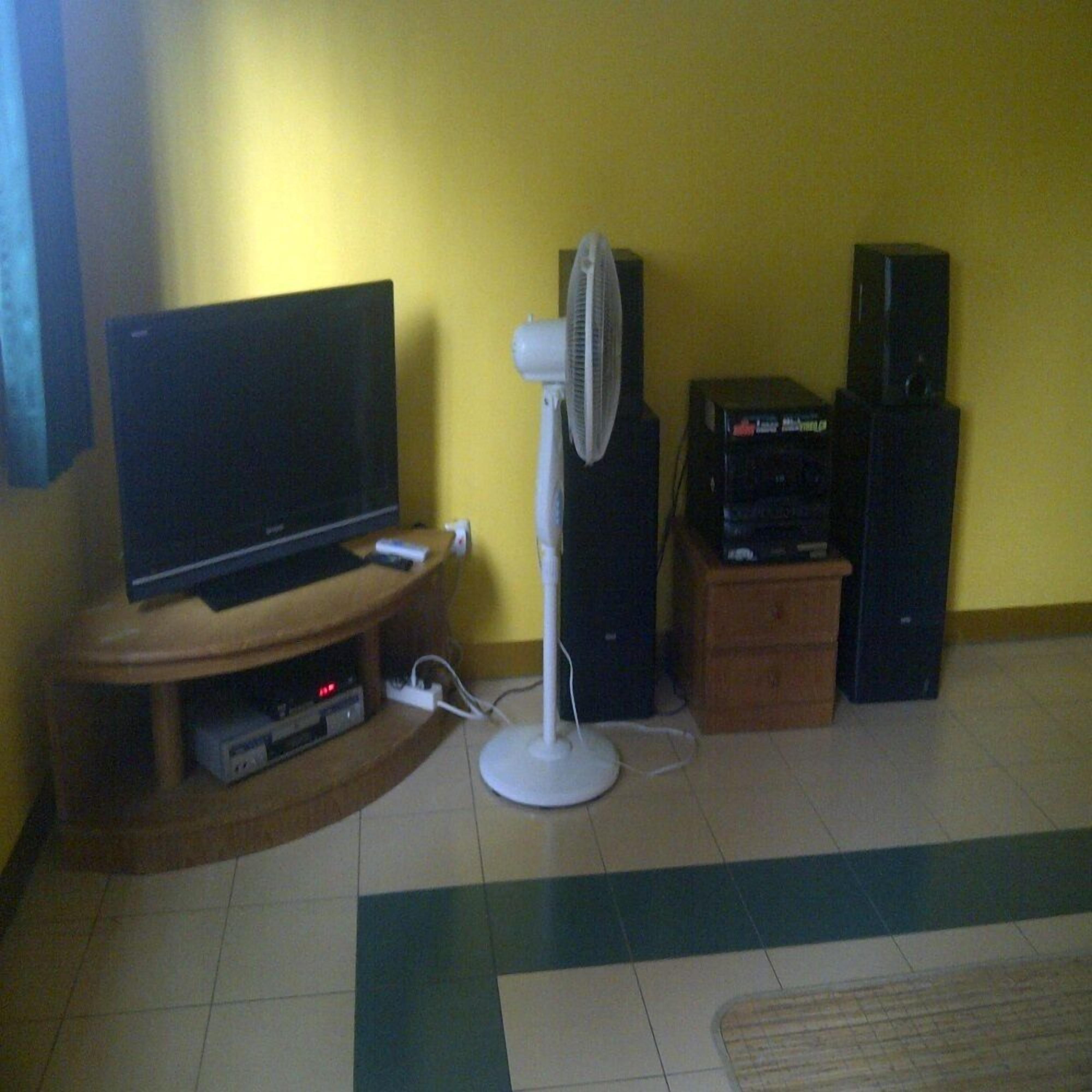 A living room with a TV on a wooden stand, a white standing fan, and two black speakers on a wooden cabinet.