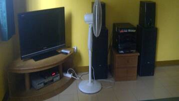 A living room with a TV on a wooden stand, a white standing fan, and two black speakers on a wooden cabinet.