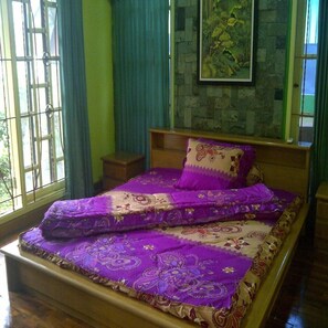 A bedroom with a bed covered in a purple and beige patterned bedspread, a wooden headboard, and a small bedside table.