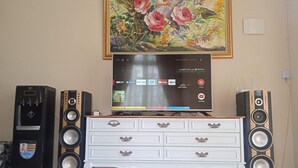 Ein Wohnzimmer mit Flachbildfernseher, zwei Lautsprechern, einem Wasserspender und einer weißen Kommode.