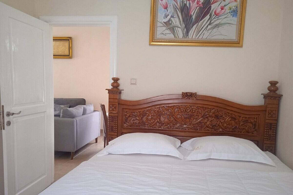 Un dormitorio con cabecera de madera, cama con ropa blanca, sofá gris y un cuadro con flores en la pared.