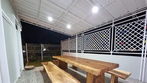 A covered outdoor area with a wooden table and benches, a white fence, and a metal gate. - Villa Kota Bunga Anyelir (Cipanas)