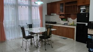 A dining area with a white table and four chairs, a kitchen with wooden cabinets and appliances, and a window with sheer curtains. - Villa Kota Bunga Anyelir (Cipanas)