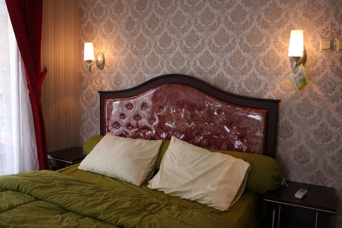 A neatly made bed with a green comforter and two pillows, a wooden headboard, and two wall-mounted light fixtures.