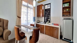 A compact kitchen with a dining table, chairs, a refrigerator, and a window with curtains. - Villa Kota Bunga Allamanda (Cipanas)