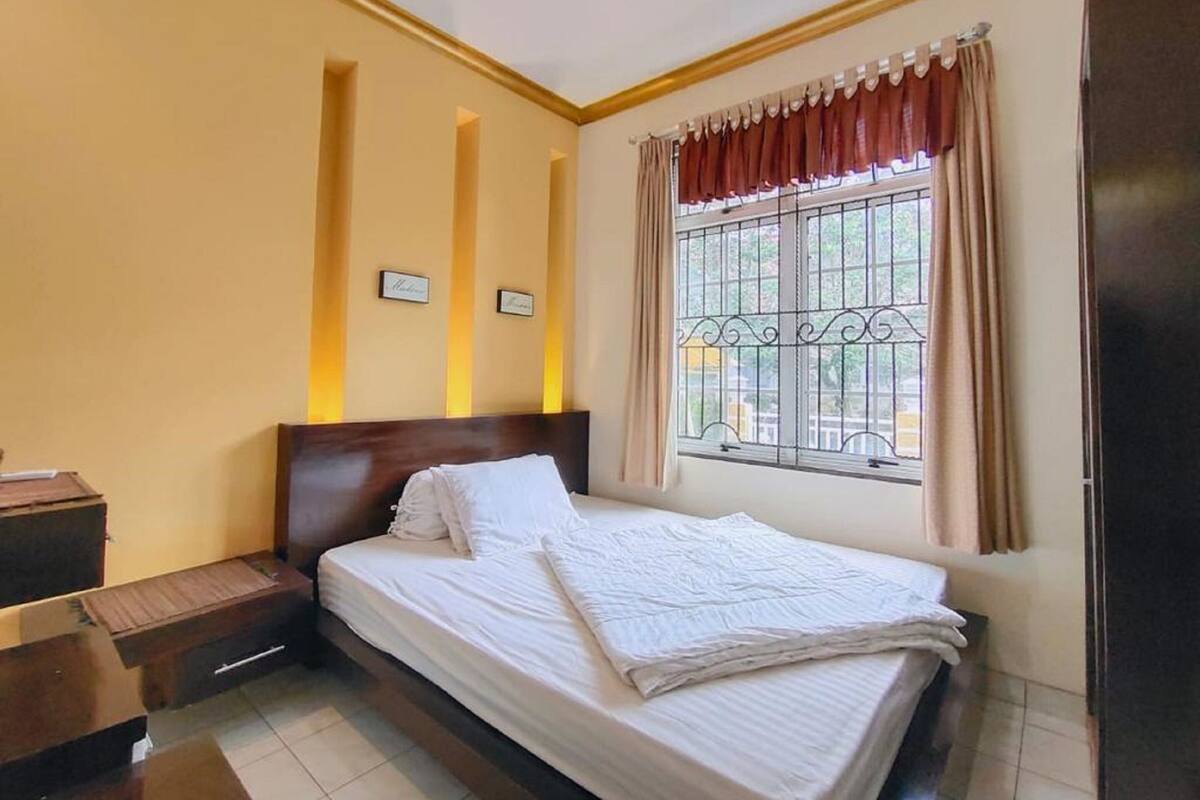 A neatly made bed in a room with a window, a bedside table, and a small cabinet.
