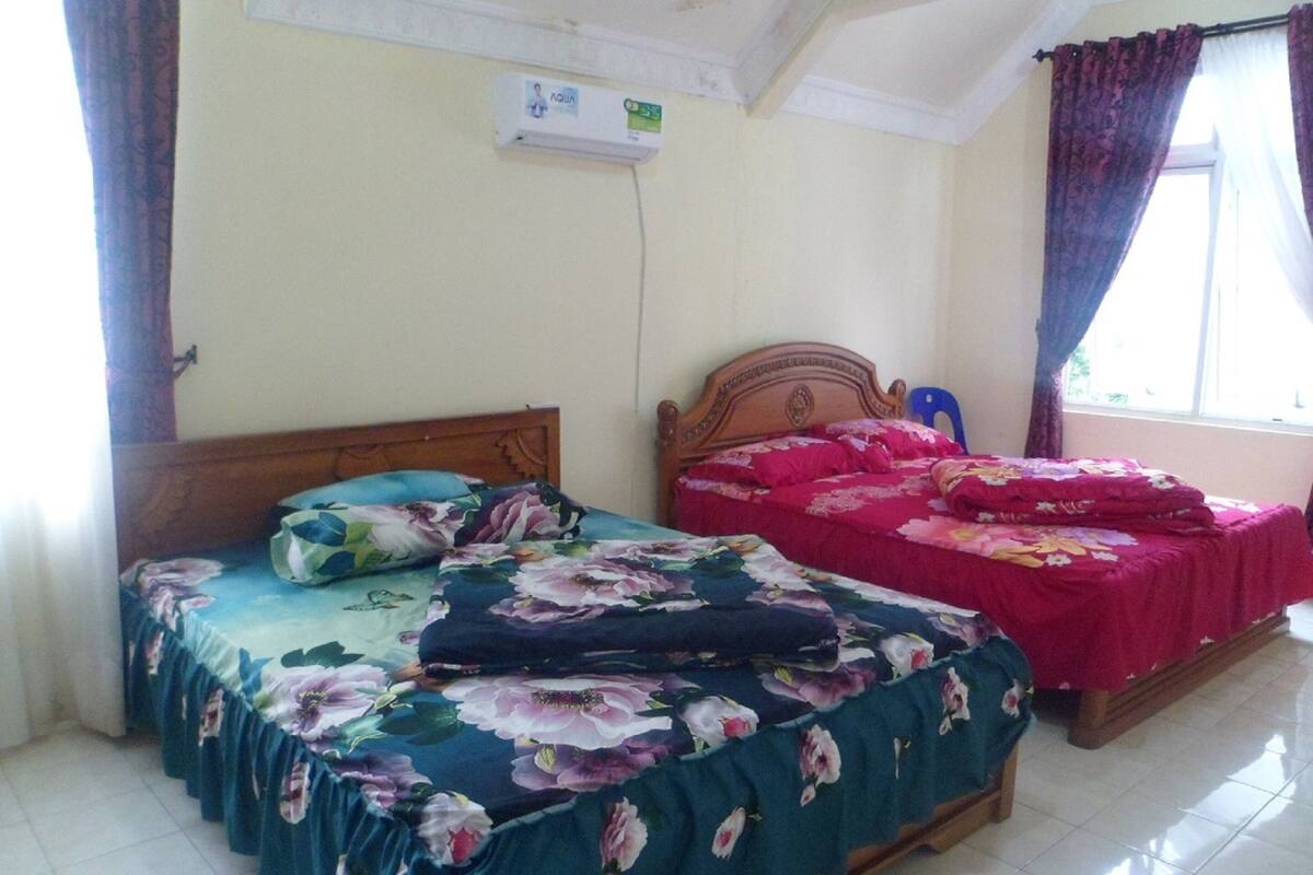 A bedroom with two beds, a wooden headboard, a window with curtains, and a wall-mounted air conditioner.