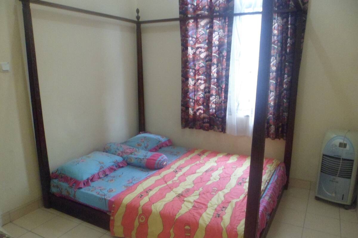 A four-poster bed with floral bedding, a window with patterned curtains, and a standing fan.