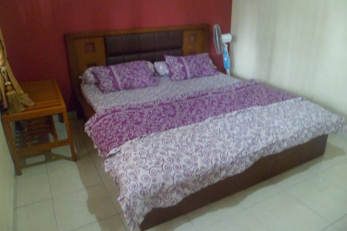 A bedroom with a large bed, a wooden nightstand, a fan, and a red wall.