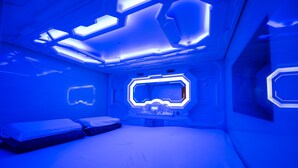 Ein futuristisches Zimmer mit einer zentralen Bedienkonsole, einem Bett mit Kissen und einem an der Wand montierten Bildschirm.