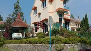 A multi-story house with a balcony, surrounded by lush greenery and a stone wall. - Villa Private Puncak (Cipanas)